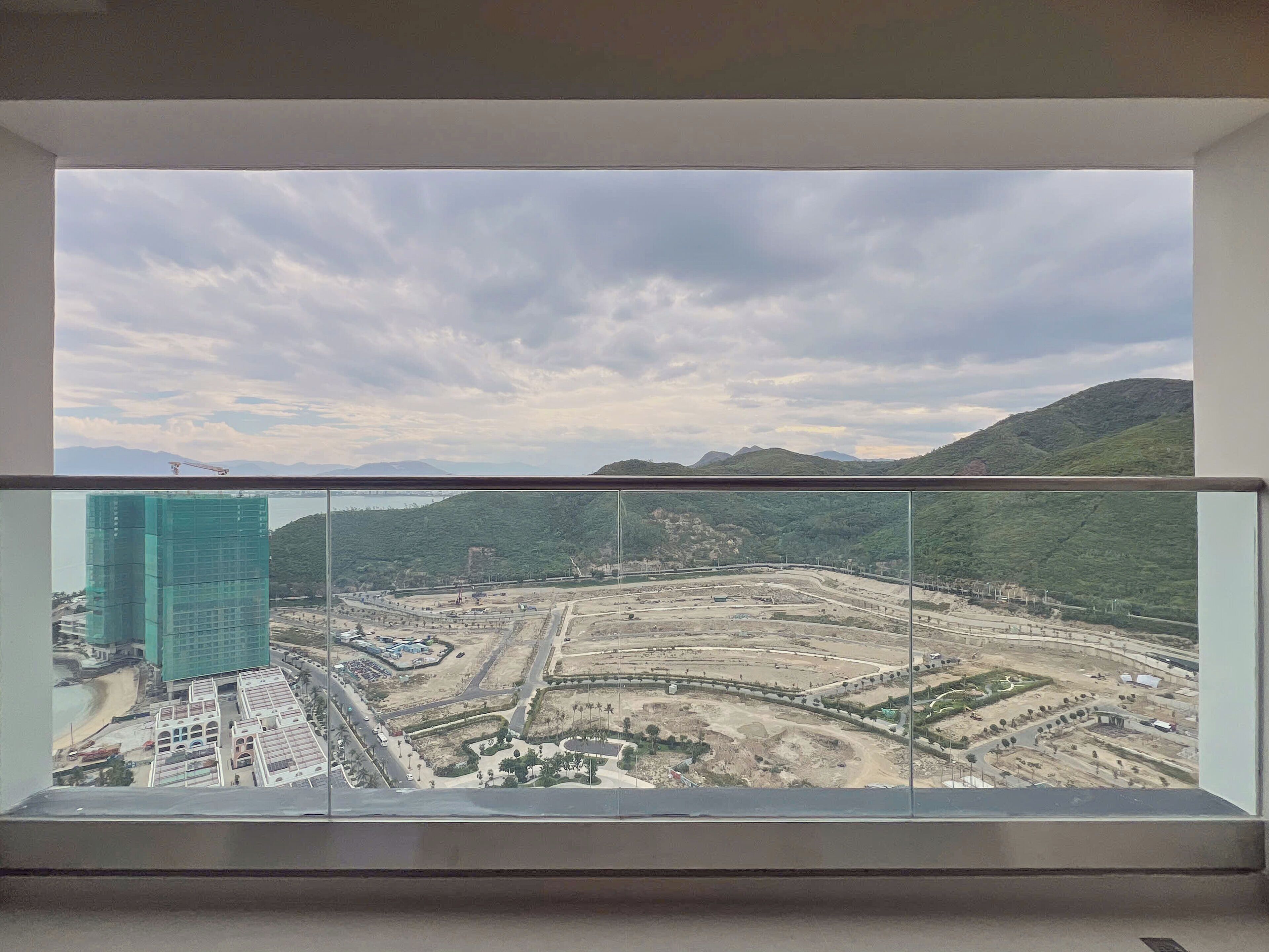 A view from a balcony with a glass railing, overlooking a construction site with a large building in the foreground and mountains in the background.