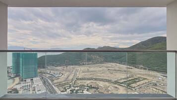 A view from a balcony with a glass railing, overlooking a construction site with a large building in the foreground and mountains in the background.
