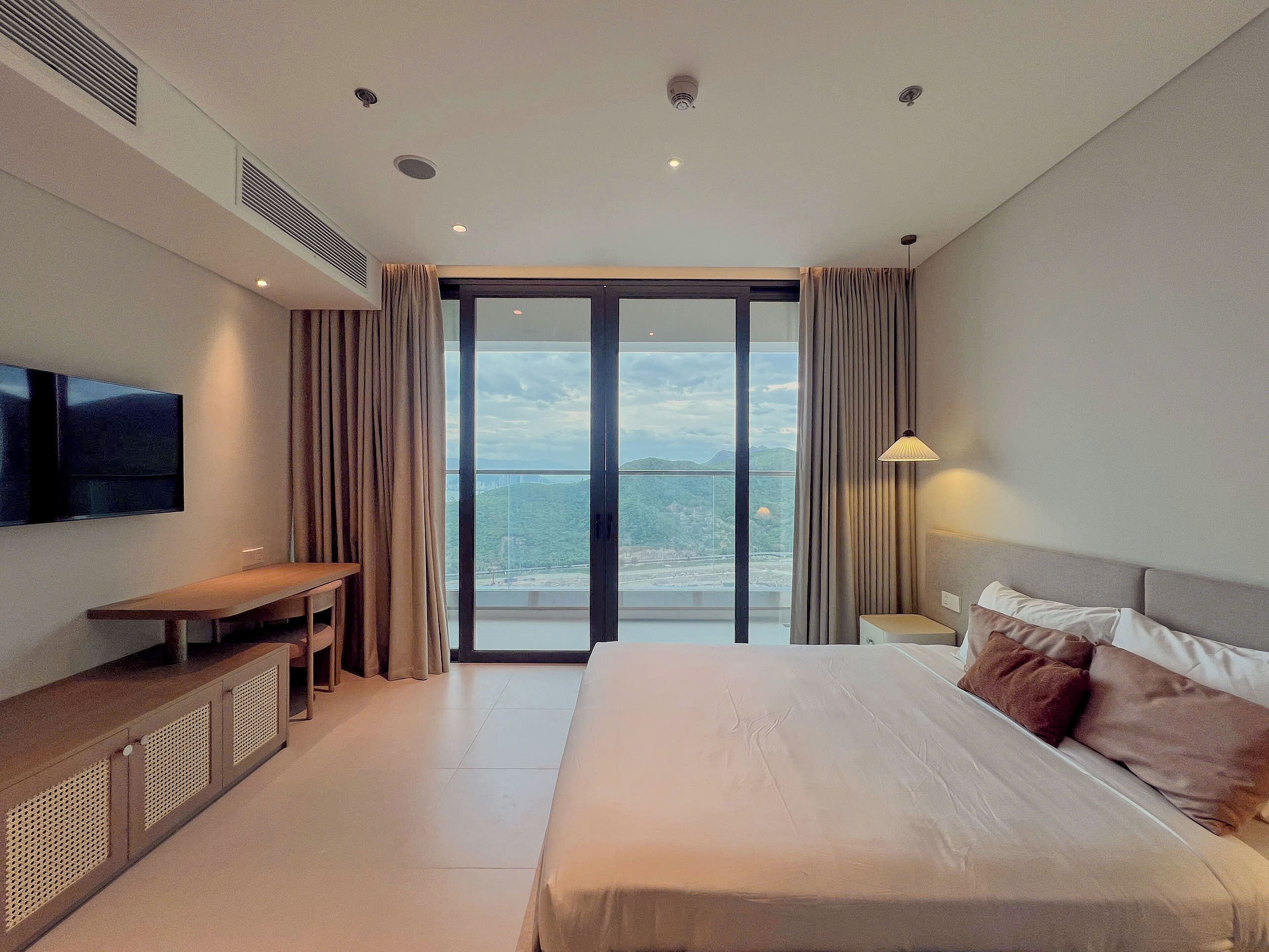 A modern hotel room with a large bed, a flat-screen TV, a desk, and a view of a cityscape through the window.