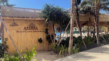 Un panneau en bois sur lequel on peut lire « ALOHA Beach Club & Restaurant », entouré de palmiers et surplombant un ciel dégagé.