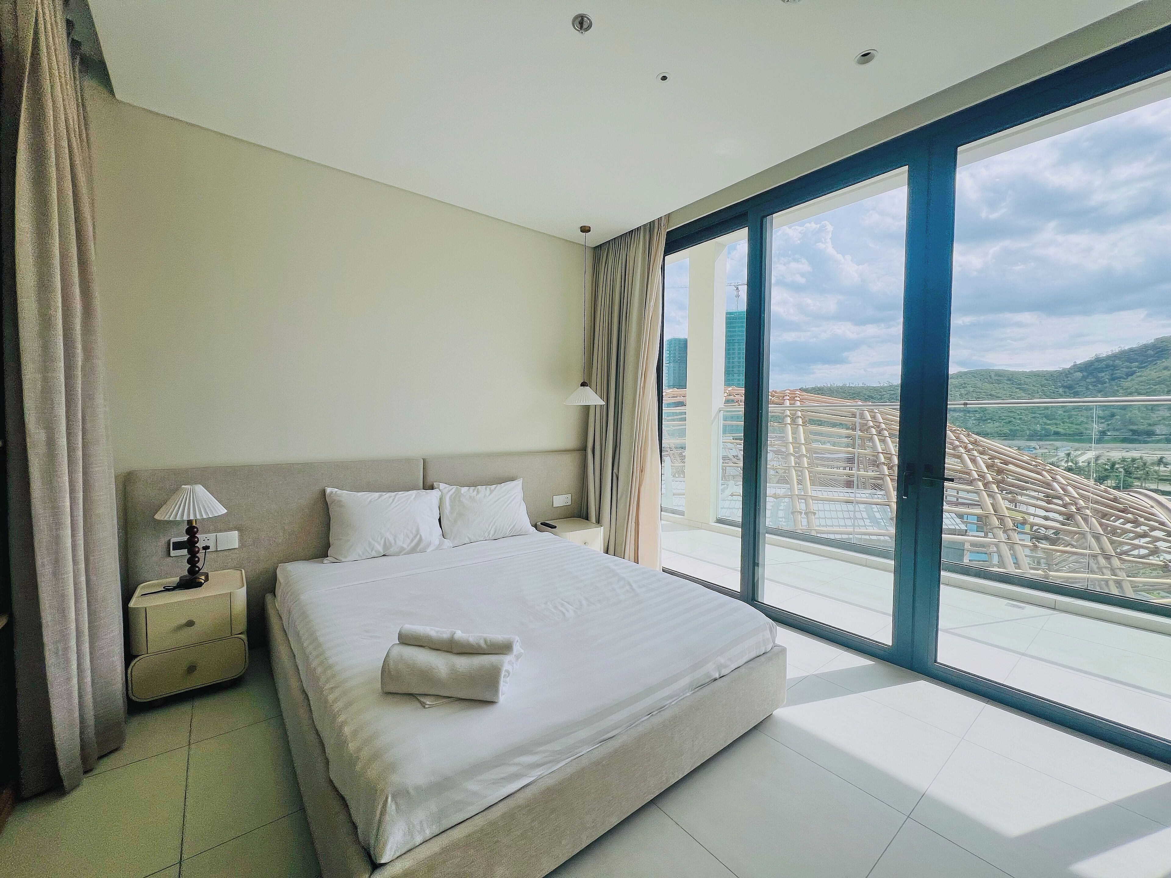A hotel room with a large bed, a nightstand, a lamp, and a view of a cityscape through a large window.