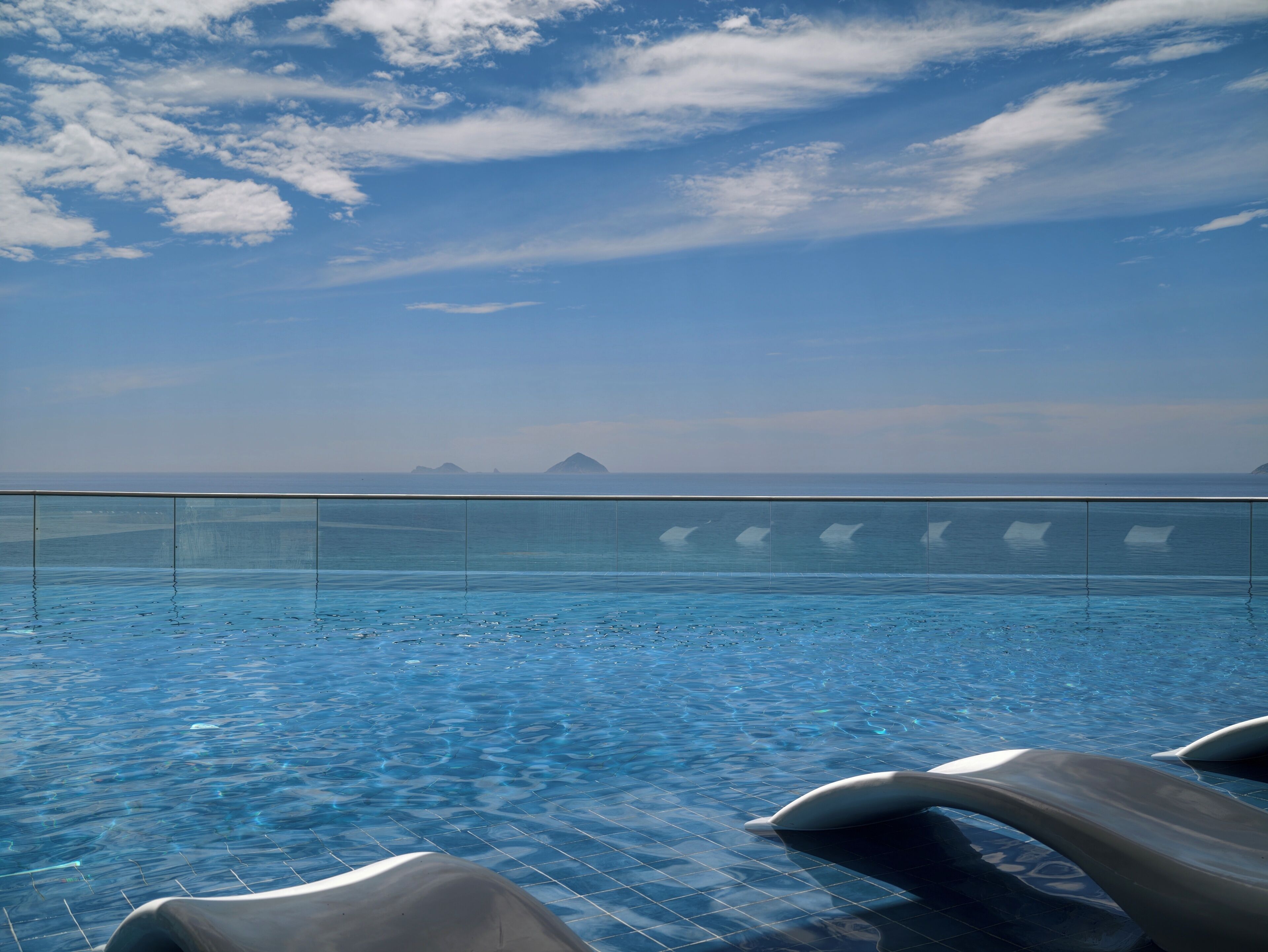 Cielo azul despejado con nubes, piscina con agua cristalina y vistas al mar y a islas distantes.