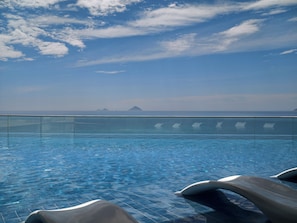 Cielo azul despejado con nubes, piscina con agua cristalina y vistas al mar y a islas distantes.