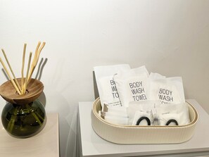 Ein Zimmer mit einer Holtschale, die Schilfhalme, eine Glasvase und eine Ablage mit weißen Handtüchern mit den Aufschriften 'BODY WASH TOWEL' und 'COTTON SET' enthält.