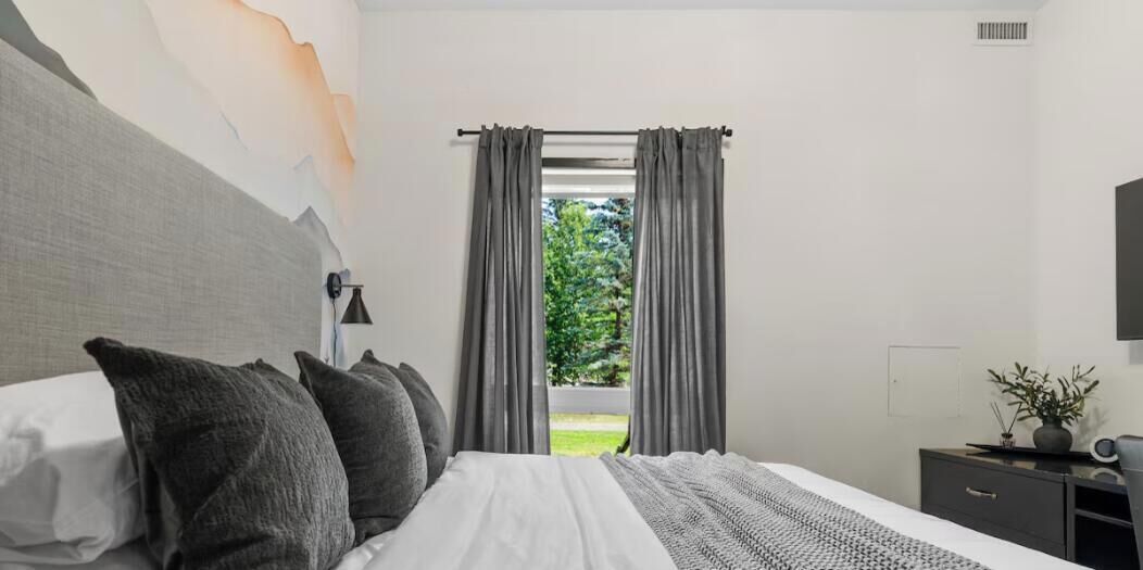 Une chambre à coucher avec un lit, une table de chevet, une lampe, une fenêtre avec des rideaux et une vue sur la verdure à l’extérieur.