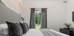 Ein Schlafzimmer mit einem Bett, einem Nachttisch, einer Lampe, einem Fenster mit Vorhängen und Blick ins Grüne.