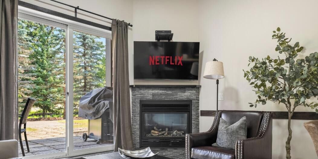 A living room with a fireplace, a television mounted above it, and a view of a forest through a sliding glass door.