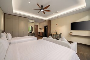 Une chambre d’hôtel avec deux lits, un ventilateur de plafond, un téléviseur fixé au mur et, au fond, une petite cuisine.