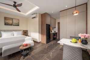 A hotel room with a bed, a small table with fruit, a chair, a kitchenette, and a ceiling fan. - Eli Living Apartment (Da Nang)