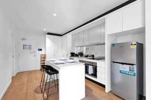 A modern kitchen with white cabinetry, a central island with bar stools, a built-in oven, and a stainless steel refrigerator. - Elegant Melbourne CBD Getaway - Pool & Gym Access (Melbourne)