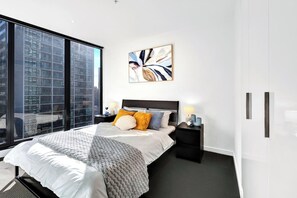 Ein modernes Hotelzimmer mit großem Fenster, einem Bett mit weißer Tagesdecke, mehreren Kissen, einem Nachttisch mit Lampe und einem an der Wand hängenden abstrakten Gemälde.
