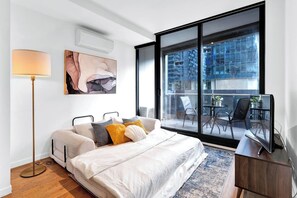 Un dormitorio moderno con cama, lámpara de noche, obra de arte y vistas de la ciudad a través de amplias ventanas.