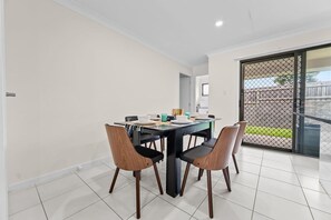 Uma área de jantar com uma mesa posta para quatro pessoas, cadeiras e vista para uma área externa através de uma porta de vidro.