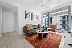 Un salon moderne avec un canapé vert, une table basse et un tapis marron. Les murs sont ornés de tableaux encadrés et un ventilateur de plafond est installé au centre.