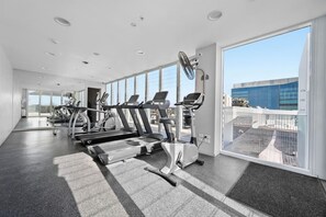 Une salle de sport moderne équipée de tapis de course, de vélos elliptiques et d’un ventilateur, avec de grandes fenêtres offrant une vue sur un immeuble.