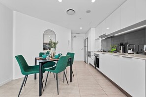 A modern kitchen with white cabinetry, a dining area with green chairs, and a large window. - Valley Stay Stylish Apartment |Parking |Pool & Gym (Newstead)