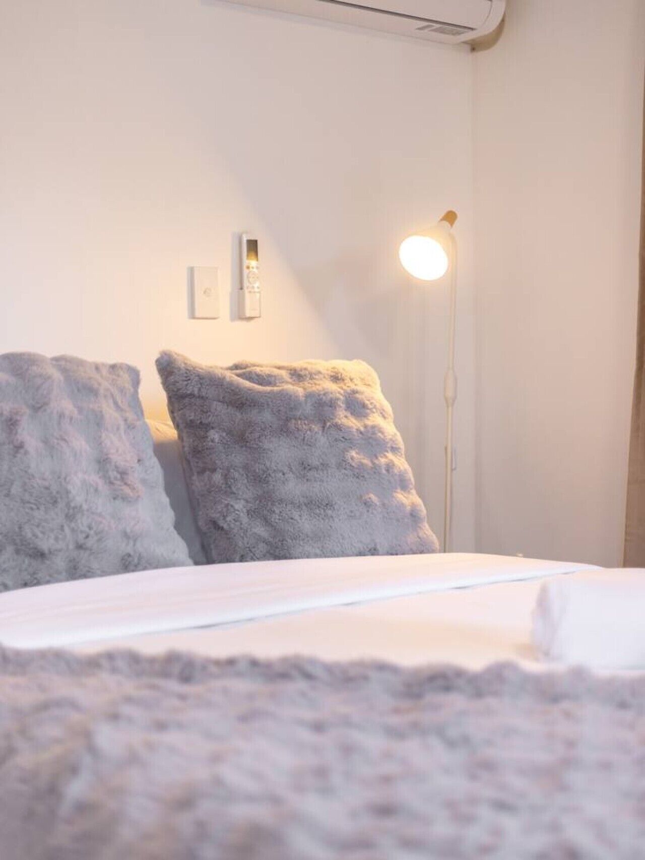 Un lit avec une literie blanche, deux oreillers gris, une lampe de sol blanche et un interrupteur fixé au mur.