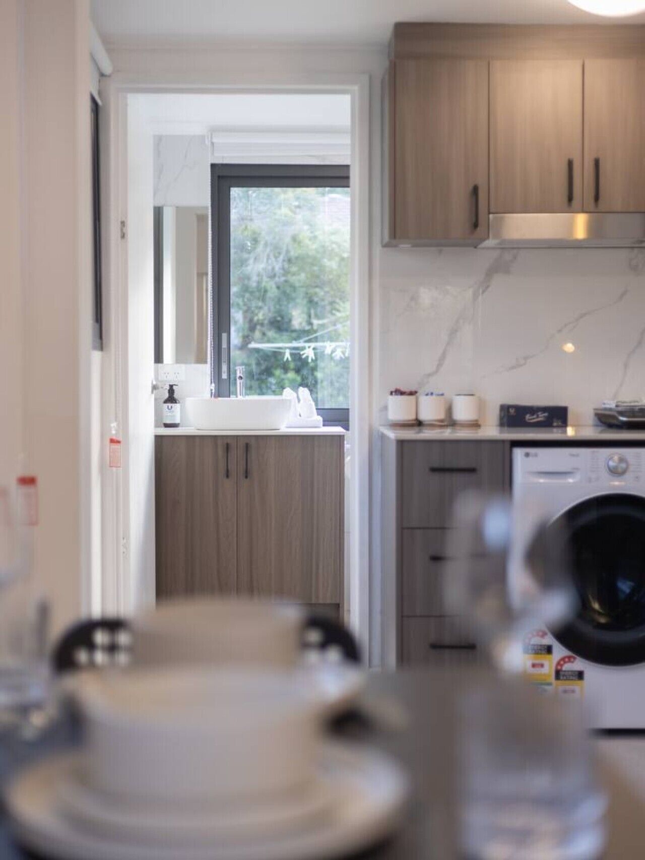 A modern kitchen with wooden cabinets, a built-in washing machine, and a window providing a view of greenery outside.