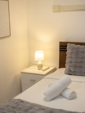 A neatly made bed with a rolled towel, a bedside lamp, and a nightstand with a phone and a remote control.