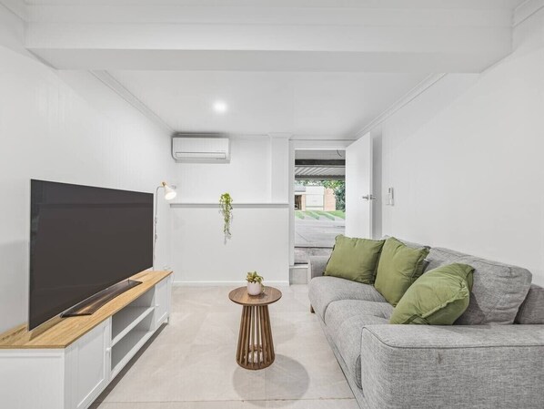 A modern living room with a flat-screen TV on a wooden stand, a grey sofa with green cushions, a small wooden side table with a plant, and a white wall with a hanging plant.