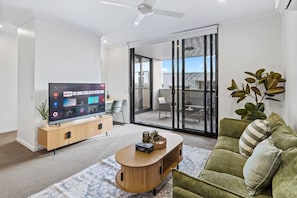 A modern living room with a flat-screen TV, a wooden coffee table, a green sofa, and a sliding glass door leading to an outdoor area.