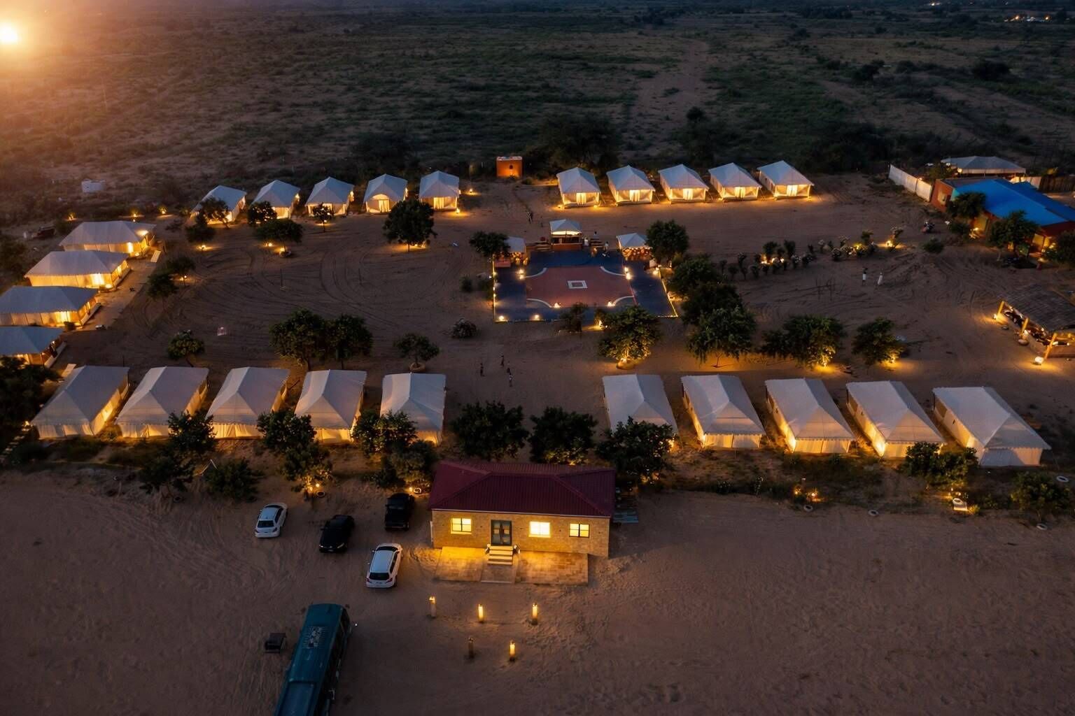 Une vue aérienne d’un campement dans le désert, avec de nombreuses tentes disposées en rangées, un bâtiment central doté d’un toit rouge et quelques véhicules garés à proximité.