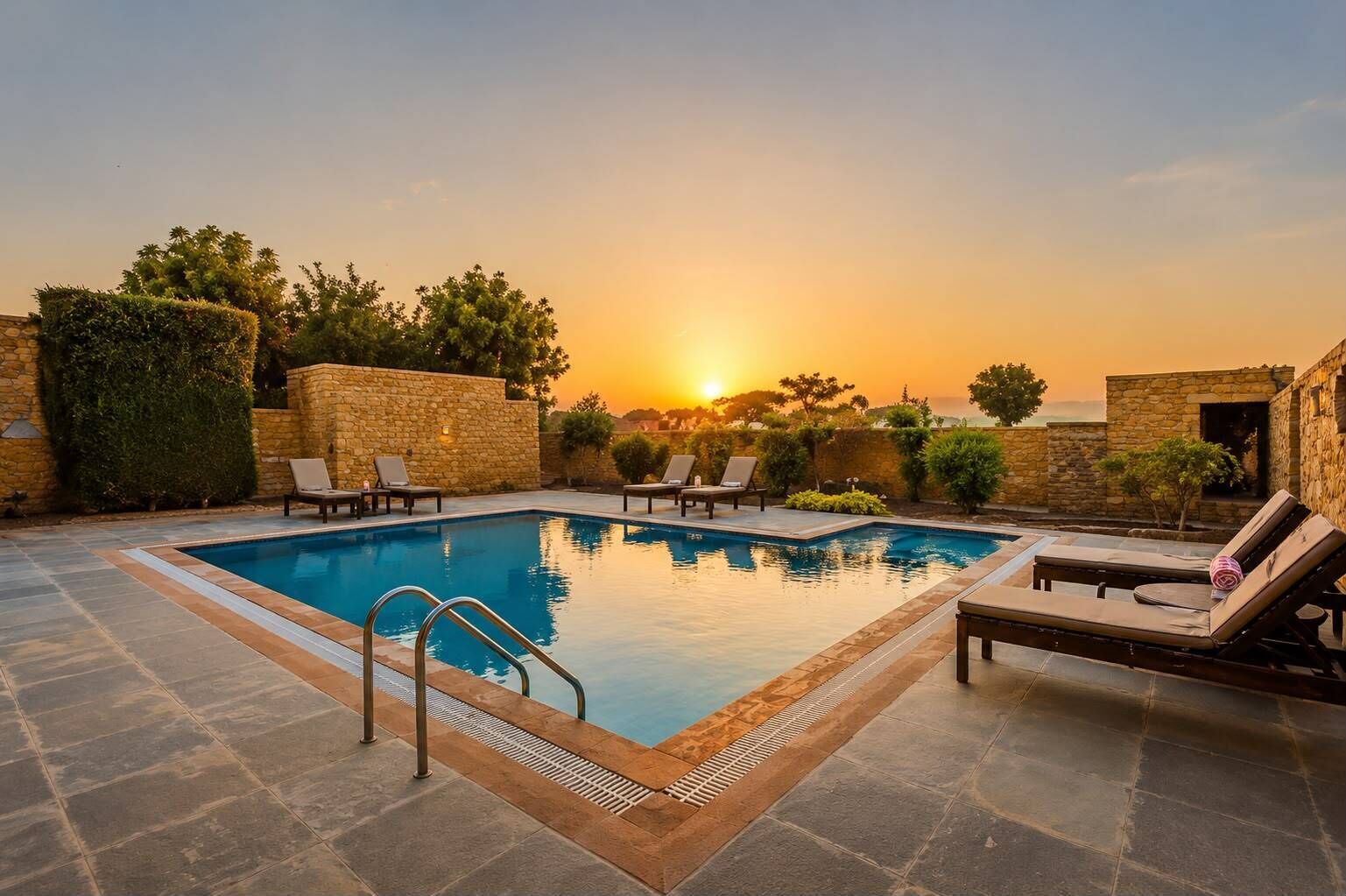 Una piscina rectangular con borde de piedra, rodeada de sillas de descanso y un muro de piedra.