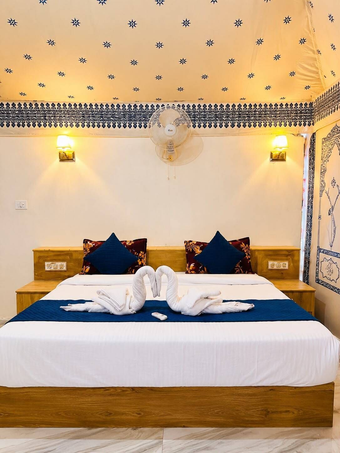 Quarto de hotel com cama, duas mesas de cabeceira, ventoinha na parede e arandelas decorativas.