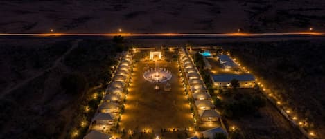 Vista aérea de un resort desértico de noche, con varias tiendas de campaña, un área de encuentro central y una piscina.