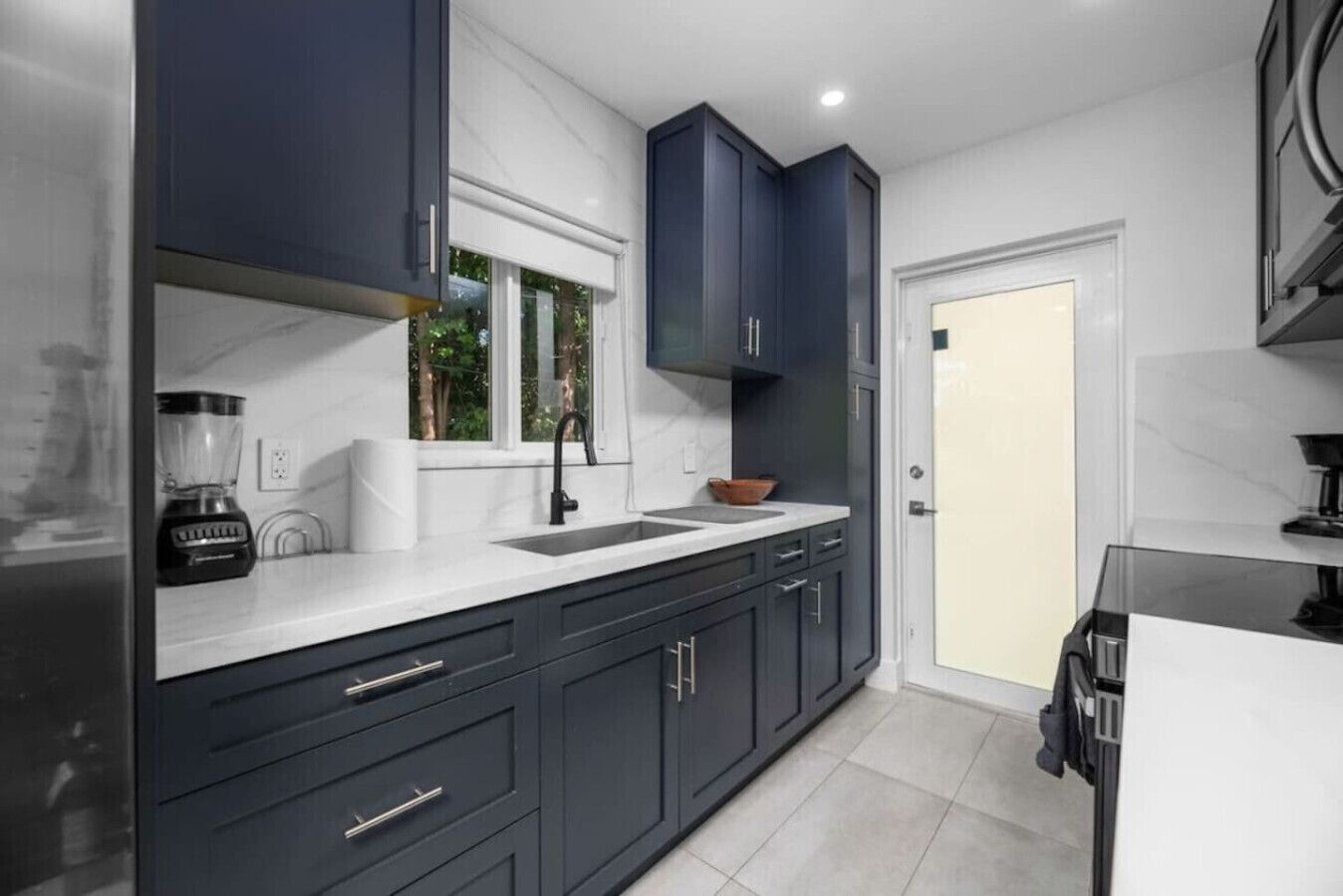 A modern kitchen with dark cabinetry, a white countertop, and a window with a view of greenery.