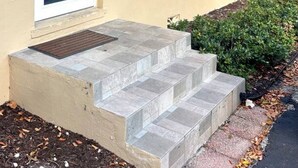 En modern byggnad med en beige fasad, en röd tegeldetalj, en vit dörr och en liten trappa av sten som leder upp till entrén.