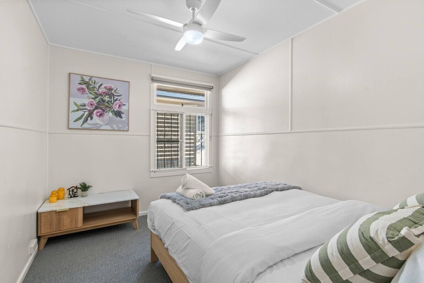 A bedroom with a bed, a ceiling fan, a window with blinds, a bedside table with a lamp, and a wall-mounted painting.