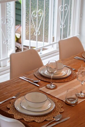 Ein Esstisch mit Tellern, Besteck und Gläsern, mit Blick auf einen Garten durch ein Fenster mit schmiedeeisernen Geländern.