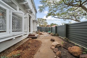 Ein Garten mit befestigtem Weg, einer kleinen Pflanzenauslage mit Steinen und einer grauen Umzäunung.