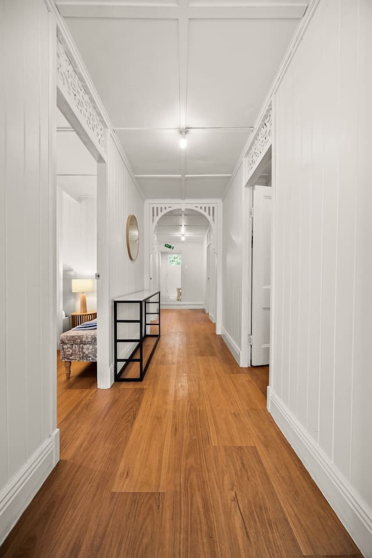 A hallway with white wainscoting, a wooden floor, and a view into a room with a bed and a lamp.