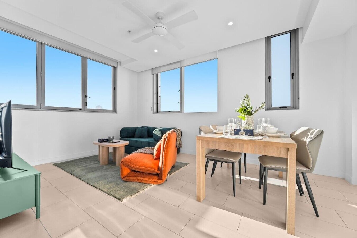A modern living room with a sofa, a dining table with chairs, a TV, and large windows.