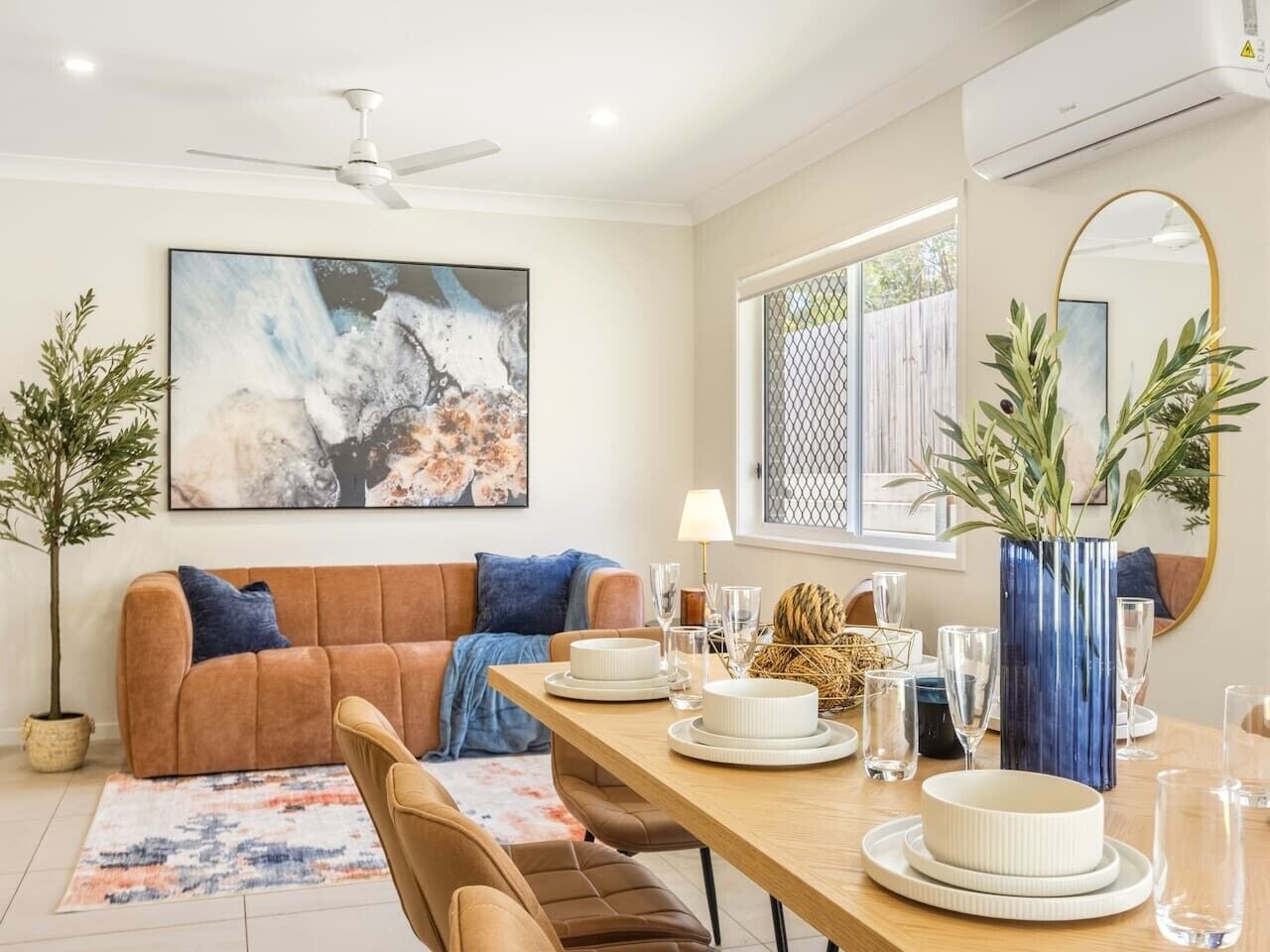 Uma sala de estar moderna com um sofá, mesa de jantar posta com pratos e copos, um grande quadro abstrato e um vaso de planta.