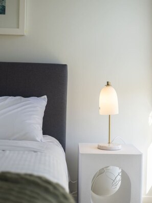 Ein ordentlich gemachtes Bett mit weißer Bettwäsche, einem dunklen Kopfteil und einem Nachttisch mit Lampe.