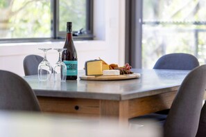 A table with a bottle of wine, two glasses, a cheese board with various cheeses and grapes, and a window with a view of greenery. - Rose Creek Chalet: Cosy 2BR w/ Resort Facilities, Crackenback Resort NSW (Crackenback)