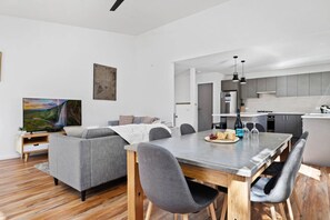 A modern living room with a grey sofa, a dining table with chairs, and a kitchen in the background. - Rose Creek Chalet: Cosy 2BR w/ Resort Facilities, Crackenback Resort NSW (Crackenback)