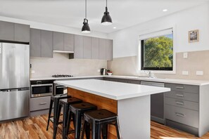 A modern kitchen with a central island, bar stools, stainless steel appliances, and a window with a view of trees. - Rose Creek Chalet: Cosy 2BR w/ Resort Facilities, Crackenback Resort NSW (Crackenback)
