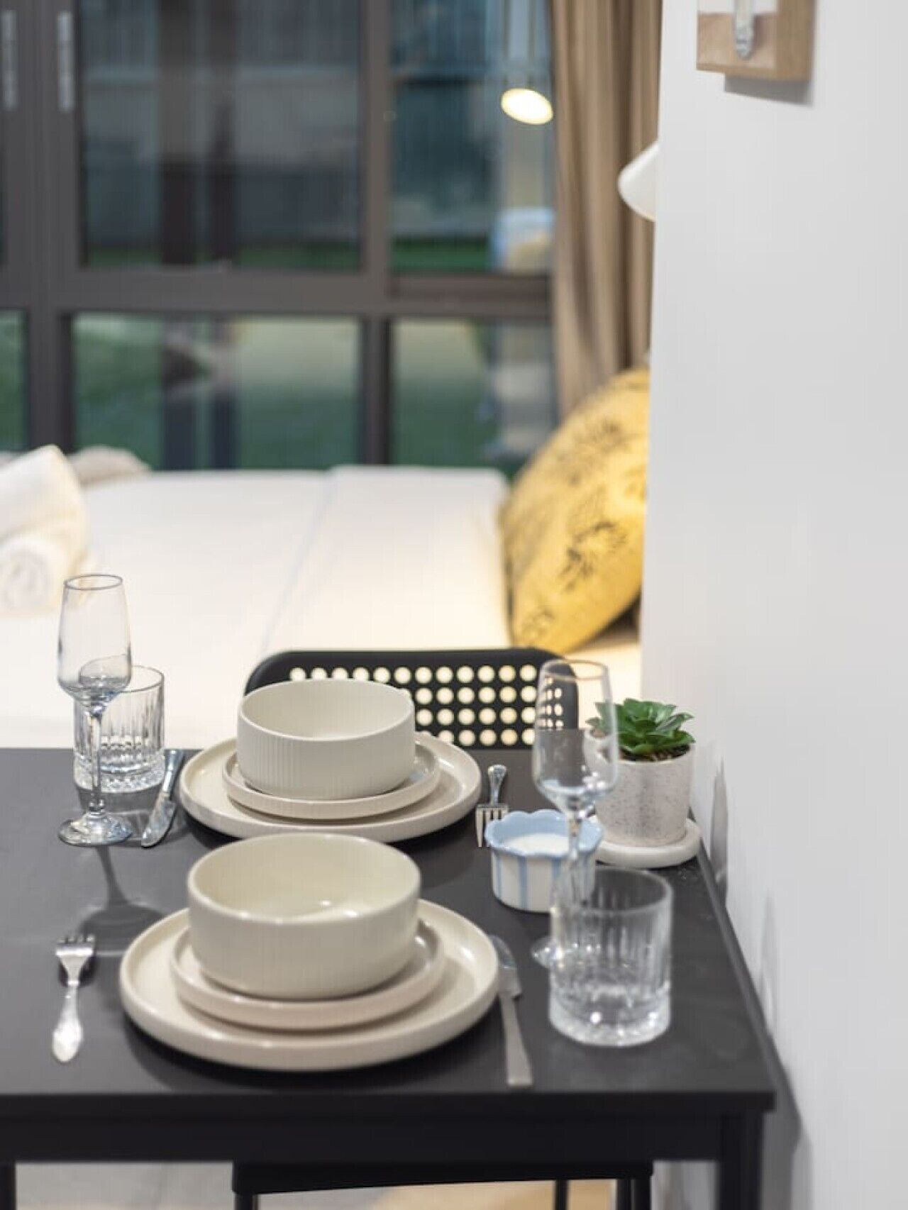 A neatly arranged dining table with plates, glasses, and cutlery, set against a modern interior with a bed and window in the background.