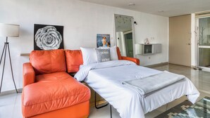 A modern hotel room with a sectional sofa, a bed with white bedding, a large mirror, and artwork on the walls.