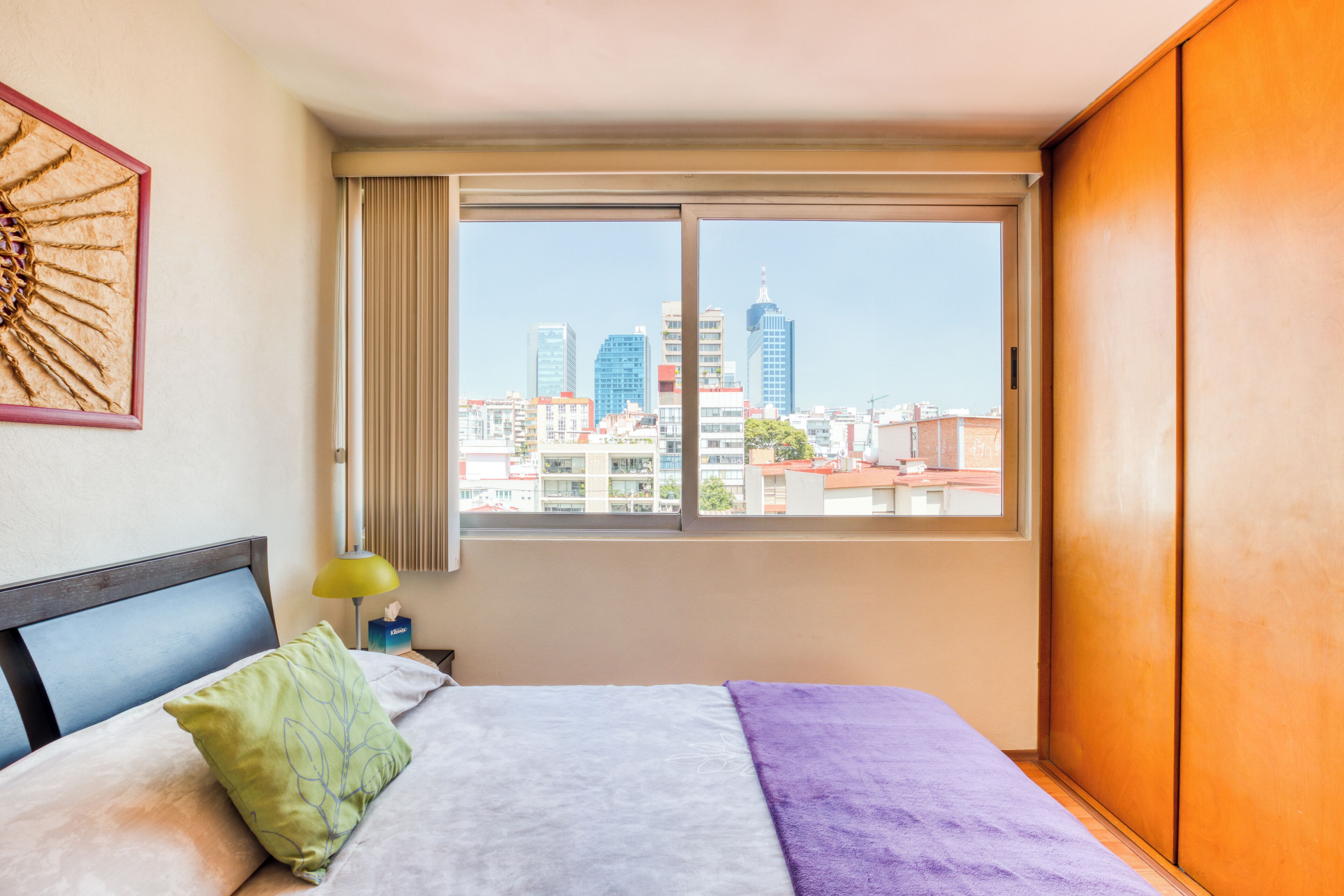 Ein Hotelzimmer mit Bett, Fenster mit Stadtblick, einer Nachttischlampe und einem gerahmten Kunstwerk an der Wand.