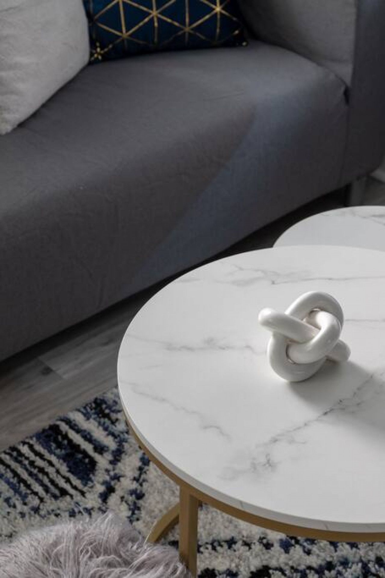 A round white marble table with a white object on it, a grey sofa with a patterned cushion, and a patterned rug.