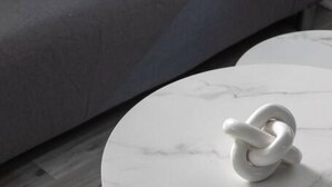 A round white marble table with a white object on it, a grey sofa with a patterned cushion, and a patterned rug.