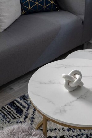A round white marble table with a white object on it, a grey sofa with a patterned cushion, and a patterned rug.
