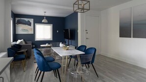 A modern living room with a dining area, featuring a white table, blue chairs, and a large window.