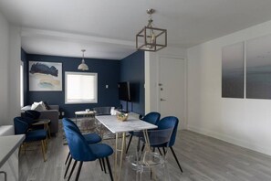 A modern living room with a dining area, featuring a white table, blue chairs, and a large window.