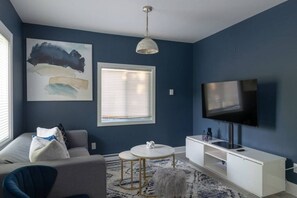 A living room with a sofa, a round coffee table, a white console table, a flat-screen TV mounted on the wall, and a painting above the sofa.